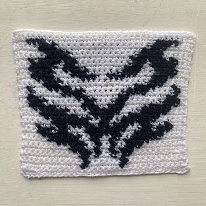 May include: A square, white crocheted item with a black, wing-like design in the center. The design is symmetrical and detailed, with the black sections resembling feathers or stylized wings. The item is handmade and has a textured surface.