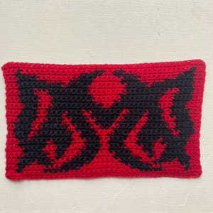 May include: A rectangular, crocheted item with a red background and a black graphic design. The design appears to be a stylized symbol or emblem. The item is handmade and has a textured appearance.