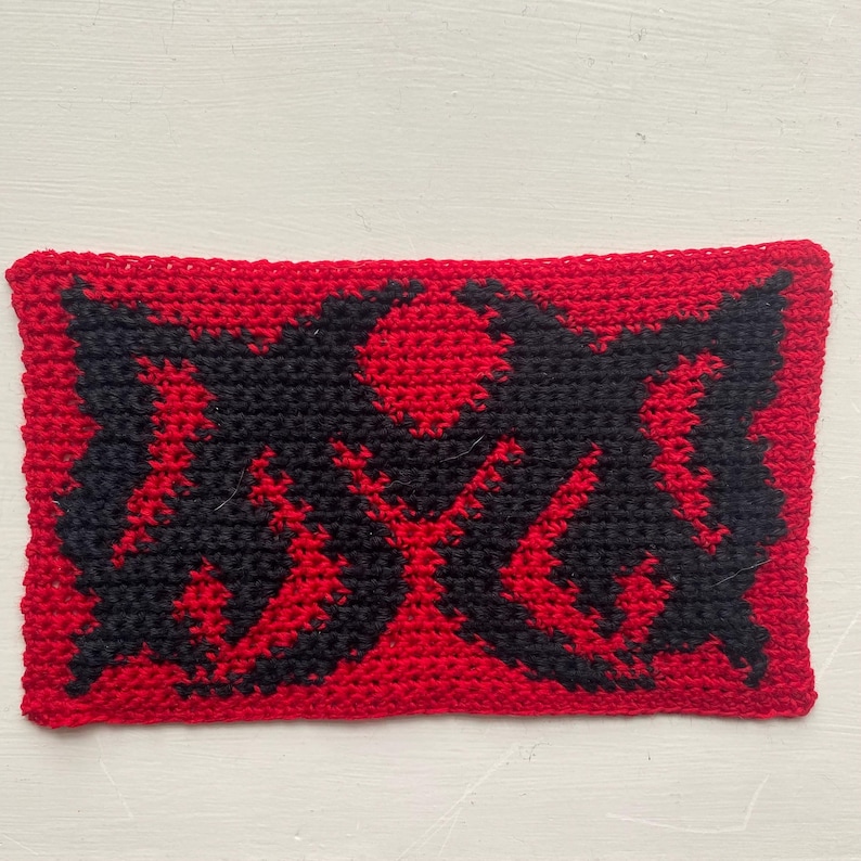 May include: A rectangular, crocheted item with a red background and a black graphic design. The design appears to be a stylized symbol or emblem. The item is handmade and has a textured appearance.
