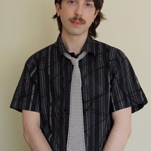 May include: A light gray, crocheted tie is the focal point of this image. The tie is worn over a black button-down shirt with a white, abstract pattern. The person in the image is wearing the tie.