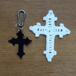 May include: Two crochet crosses on a wooden surface. One is white, the other is black and attached to a silver keychain. The crosses have a decorative, ornate design. The white cross is larger than the black cross.