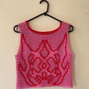 Op de afbeelding: Een roze gehaakte top met een rood decoratief ontwerp en afwerking. De mouwloze top hangt aan een zwarte hanger tegen een neutrale achtergrond. Het haakpatroon is open en luchtig.