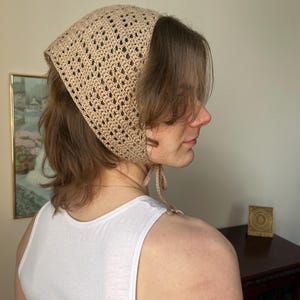 Puede incluir: Un pañuelo de ganchillo beige con forma cuadrada y diseño calado. El cubrecabezas se ata en la nuca con lazos beige a juego. El pañuelo se lleva sobre el cabello.
