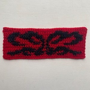 May include: A rectangular, hand-crocheted headband in vibrant red, featuring a symmetrical black design. The black pattern appears to be an abstract floral or stylized shape. The headband is made of a textured yarn, suggesting a handmade craft item.