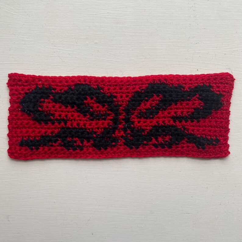May include: A rectangular, hand-crocheted headband in vibrant red, featuring a symmetrical black design. The black pattern appears to be an abstract floral or stylized shape. The headband is made of a textured yarn, suggesting a handmade craft item.
