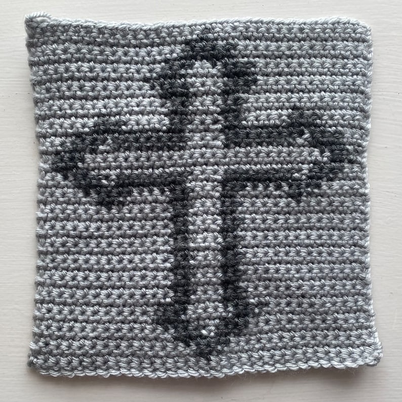May include: A gray crocheted square with a black cross design in the center.