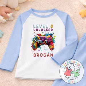 May include: White and pale blue long-sleeved pajama top featuring a vibrant video game controller design. The text "LEVEL 8 UNLOCKED" and a name are printed on the shirt. The pajama top has pale blue sleeves.