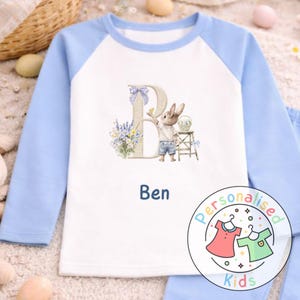 May include: A white and light blue long-sleeved shirt with a cartoon bunny and the name "Ben". The shirt features a large letter "B" with a bunny illustration, flowers, and a small stool. The shirt has a round neckline.
