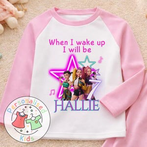 May include: Pink and white long-sleeved pajama top featuring a graphic of three cartoon singers, a star, and the name "HALLIE". The text "When I wake up I will be" is above the graphic. The pajama top has pink sleeves and a white body.