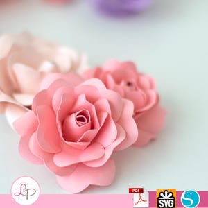 Peut inclure: Deux roses en papier rose avec des pétales superposés. Les roses sont disposées en grappe, la plus grande rose au premier plan et la plus petite rose derrière.