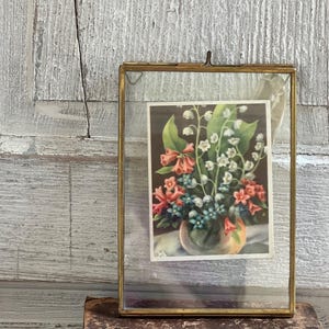 May include: A vintage brass and glass picture frame holding a floral print. The print features a bouquet of flowers in shades of pink, white, and blue. The frame is resting on a stack of old books against a weathered, white-painted wooden background.