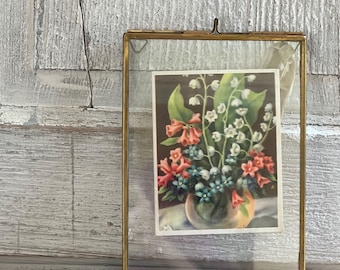 May include: A vintage brass and glass picture frame holding a floral print. The print features a bouquet of flowers in shades of pink, white, and blue. The frame is resting on a stack of old books against a weathered, white-painted wooden background.