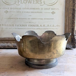 May include: A brass bowl with a scalloped rim and a circular base. The bowl has a metallic gold finish with visible signs of wear and patina. The bowl is positioned in front of a framed print featuring the text PLANT FLOWERS and other botanical details.