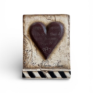 May include: A rectangular decorative piece with a dark brown heart shape at the center. The heart is embossed with text and a crown. The background is off-white with a distressed, vintage look. The base has black and white stripes.