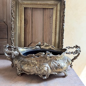 May include: An ornate brass centerpiece bowl featuring intricate relief carvings of cherubs and floral motifs, with two decorative handles on the sides and four small feet. The bowl has a dark interior and a polished metallic exterior finish.