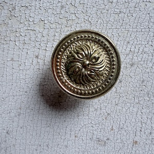 May include: A circular gold-toned metal button featuring a raised, swirling floral or sunburst pattern at the center. The design is framed by a border of small, evenly spaced raised dots along the outer edge, creating a textured metallic finish.