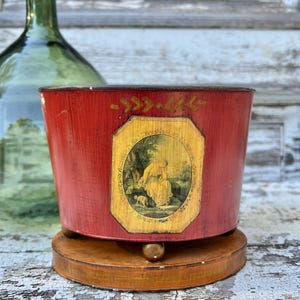 May include: A red, cylindrical container with a wooden base. The container features a framed illustration of a person and a dog. The container is likely vintage or antique, with a distressed finish.