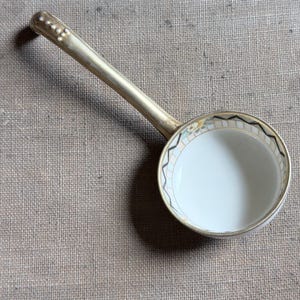 May include: A porcelain ladle with a long, metallic gold-toned handle featuring a row of small raised dots. The circular bowl has a white interior and a decorative rim with a repeating black and gold geometric pattern and floral accents.