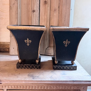 May include: Two black decorative planters with gold trim and a gold fleur-de-lis emblem. The planters have a square base with gold accents and are displayed on a wooden surface.