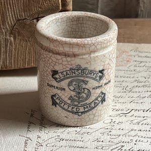 May include: A small, cylindrical, antique ceramic jar with a cracked, off-white glaze. The jar features a black and white label with the text "J. SAINSBURYS POTTED MEATS". The jar is approximately 3 inches tall and is a vintage kitchenware item.