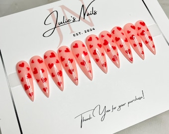 May include: Set of ten pink stiletto false nails with red heart designs. The nails are displayed on a white card with the text "Julie's Nails EST. 2024" and "Thank You for your purchase!"