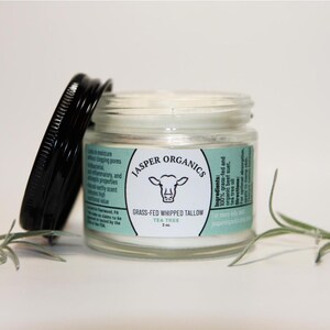 May include: A glass jar with a white lid containing a white cream. The jar has a label that says "Jasper Organics" and "Grass-Fed Whipped Tallow Tea Tree 2 oz."