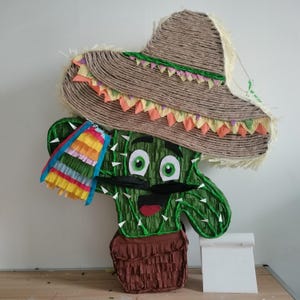 Peut inclure: Une piñata cactus verte dans un pot marron, portant un grand sombrero en corde beige avec une bordure en papier triangulaire colorée. Le cactus présente une moustache noire, des yeux blancs et verts, une bouche rouge et une frange arc-en-ciel.