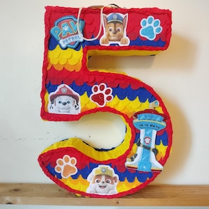 Peut inclure: Une piñata en forme de chiffre cinq décorée de franges en papier rouge, jaune et bleu. Elle présente des découpes des personnages de la Pat' Patrouille, dont Chase, Marcus et Ruben, ainsi que le logo, la tour de contrôle et des empreintes.