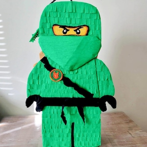 Könnte beinhalten: Eine grüne Ninja-Piñata aus Fransenpapier, mit schwarzem Gürtel, orangefarbenem Emblem und einer Maske mit gelben Augen. Die Piñata ist im Blockstil gestaltet, ähnlich einer Lego-Figur, mit einer Schlaufe zum Aufhängen.