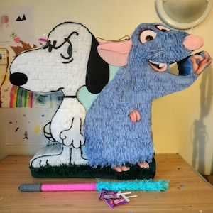 Peut inclure: Une piñata représentant un chien blanc avec une oreille et un nez noirs à côté d'un rat bleu aux grands yeux et aux oreilles roses. Les personnages sont montés sur une base verte. Un bâton rose et bleu avec un manche gris est posé devant.