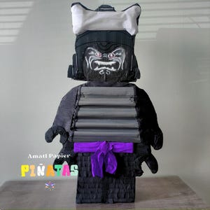 May include: A handmade piñata in the shape of a Lego-style samurai warrior. The piñata has a black body, grey armour, a white helmet, and a purple sash. The face has a detailed painted design. The text "PINATAS" is visible.
