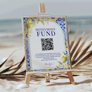Könnte beinhalten: Ein weißes Schild mit goldenem Rahmen und einem blauen und weißen Kachelbord. Das Schild trägt die Aufschrift "Honeymoon Fund" und einen QR-Code. Das Schild steht auf einem Holzstaffelei und befindet sich auf einem Strand.