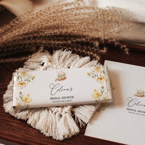 以下が含まれることがあります： 白い背景に花柄のデザインと「Celina's Bridal Shower Thanks for coming」の文字が書かれた白いチョコレートバー。チョコレートバーは白い編み物のコースターの上に置かれています。