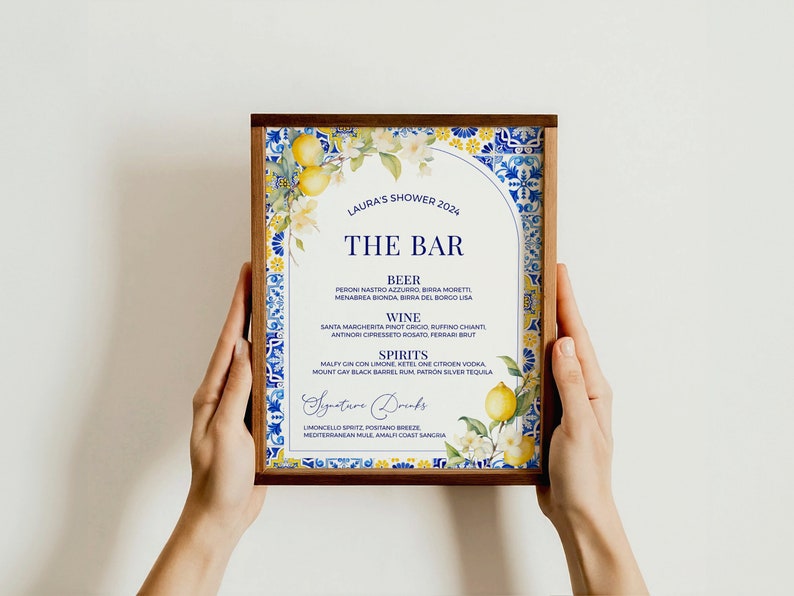 Op de afbeelding: Een ingelijst, afdrukbaar bord met een blauwe en witte betegelde rand en een citroenmotief. Het bord vermeldt de dranken die beschikbaar zijn aan een bar voor een bruidsdouche. De tekst luidt "Laura's Shower 2024", "The Bar", "Beer", "Wine", "Spirits", "Signature Drinks".