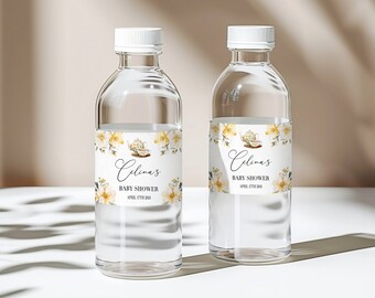 Puede incluir: Dos botellas de agua de vidrio transparente con etiquetas blancas. Las etiquetas presentan un diseño de acuarela de flores amarillas y una tetera con una taza y un platillo. El texto en las etiquetas dice "Celina's Baby Shower April 17th, 2021".