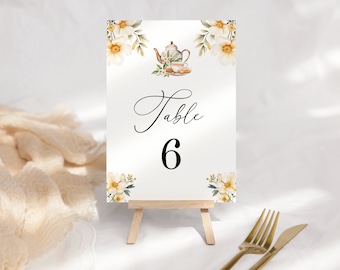 May include: White table number card with a floral design and a teapot and cup illustration. The card says "Table 6" and is displayed on a wooden easel.