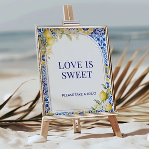 Puede incluir: Un cartel blanco con un borde azul y amarillo, en un caballete, que dice "Love is Sweet" y "Please take a treat".