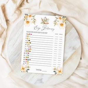 May include: A printable game card with a floral design and the title "Emoji Pictionary". The card has a list of numbers and emojis, with spaces for players to write down their guesses and whether they were right or wrong.