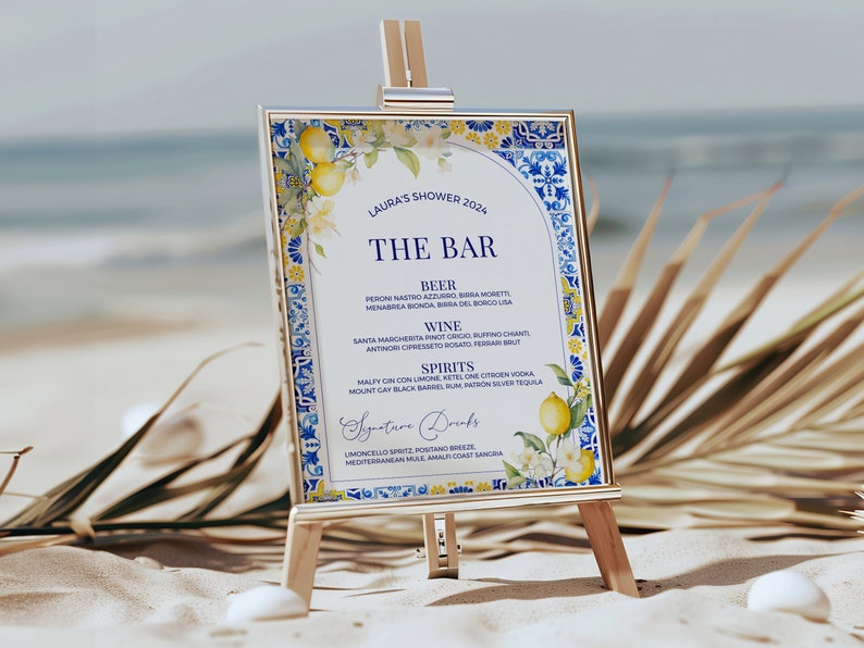 Op de afbeelding: Een bord met een blauwe en witte betegelde rand en een citroen- en bloemenontwerp. Het bord vermeldt "Laura's Shower 2024" en "The Bar". Het bord vermeldt een verscheidenheid aan dranken, waaronder bier, wijn, sterke drank en signature drinks.