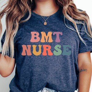 May include: A navy blue t-shirt with the text "BMT NURSE" in rainbow colors. The letters are stylized with rounded edges.