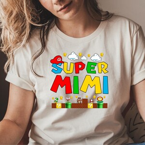 May include: White t-shirt with a colorful graphic of the words "Super Mimi" in a video game style. The graphic includes a red and white hat, clouds, coins, and a green pipe.