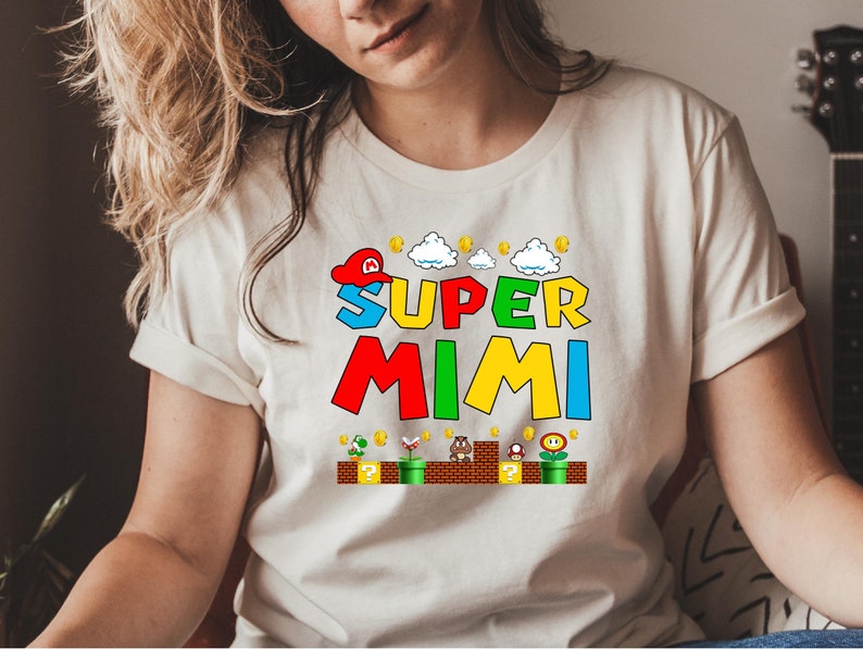 May include: White t-shirt with a colorful graphic of the words "Super Mimi" in a video game style. The graphic includes a red and white hat, clouds, coins, and a green pipe.