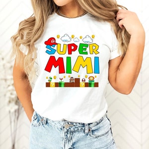 May include: White t-shirt with a colorful graphic of a Super Mario-themed design. The graphic says "SUPER MIMI" with a cartoon character wearing a red hat and a cloud above the text. The graphic also includes coins, a mushroom, a flower, and a turtle.