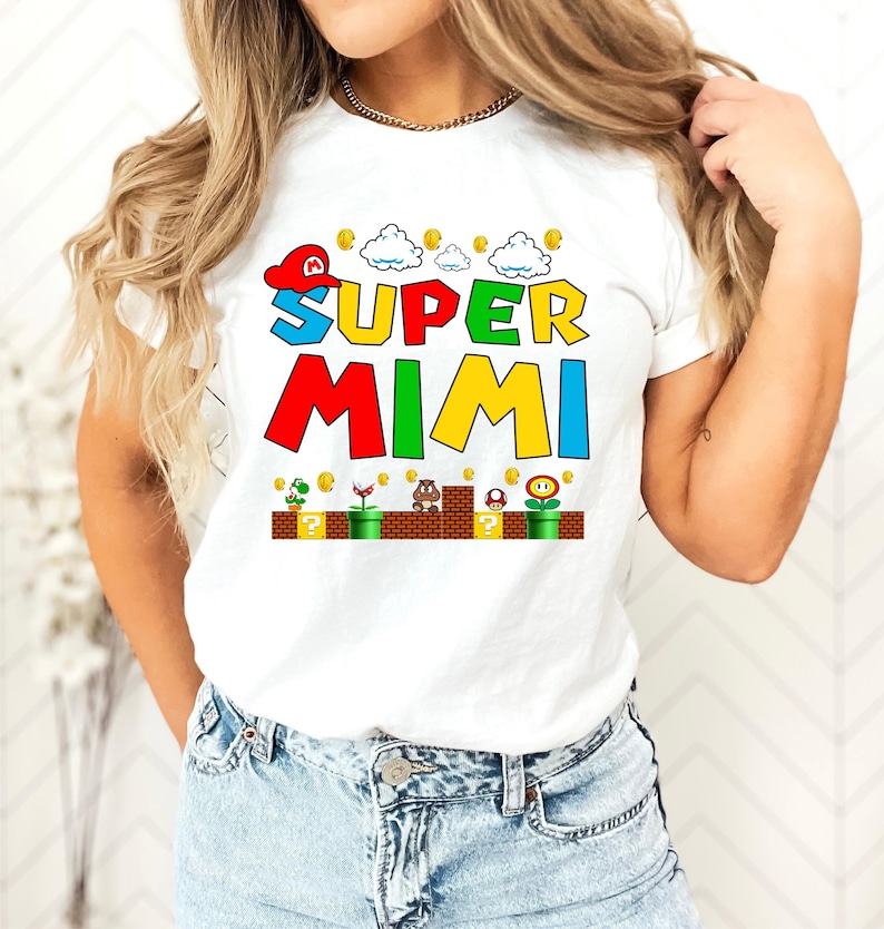 May include: White t-shirt with a colorful graphic of a Super Mario-themed design. The graphic says "SUPER MIMI" with a cartoon character wearing a red hat and a cloud above the text. The graphic also includes coins, a mushroom, a flower, and a turtle.