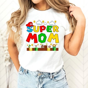 May include: White t-shirt with a colorful graphic of the words "SUPER MOM" in a video game style. The graphic includes a red and white hat, clouds, coins, and a video game level.