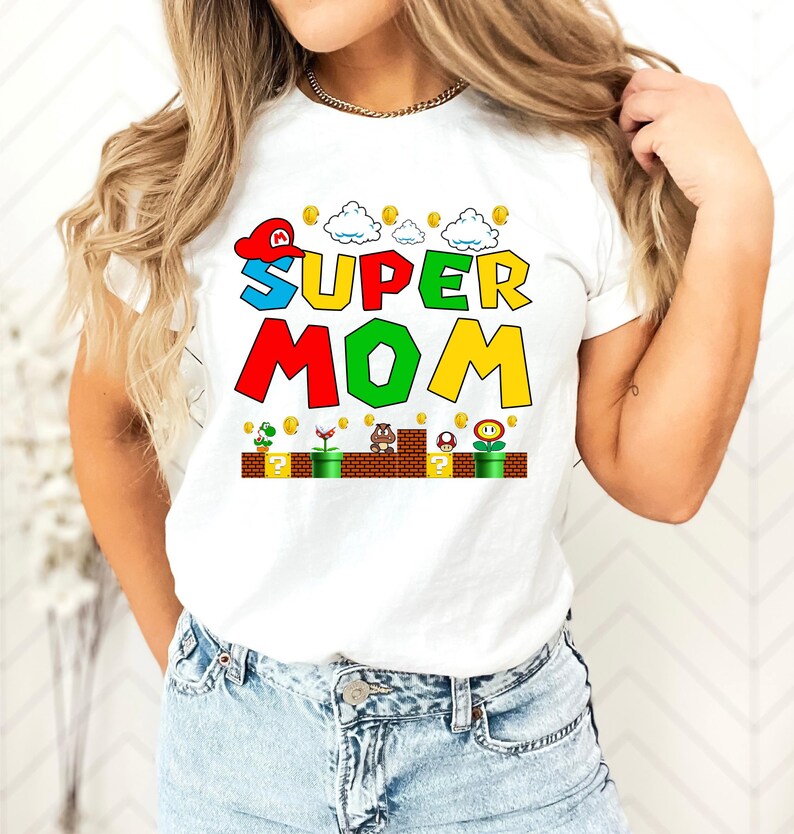 May include: White t-shirt with a colorful graphic of the words "SUPER MOM" in a video game style. The graphic includes a red and white hat, clouds, coins, and a video game level.