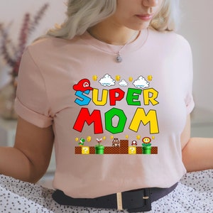 May include: A pink t-shirt with a graphic of a Super Mario-themed design that says "Super Mom" with a red, green, yellow, and blue color scheme.
