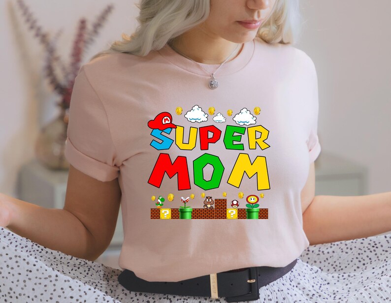May include: A pink t-shirt with a graphic of a Super Mario-themed design that says "Super Mom" with a red, green, yellow, and blue color scheme.