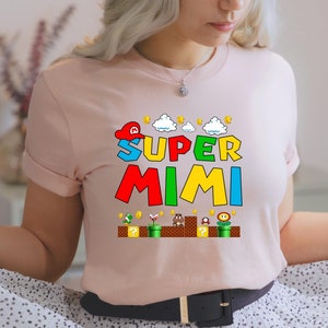 May include: A pink t-shirt with a graphic of a video game character wearing a red hat and the text "SUPER MIMI" in colorful letters. The graphic also includes a background of clouds, coins, and a brick platform with a question mark block.