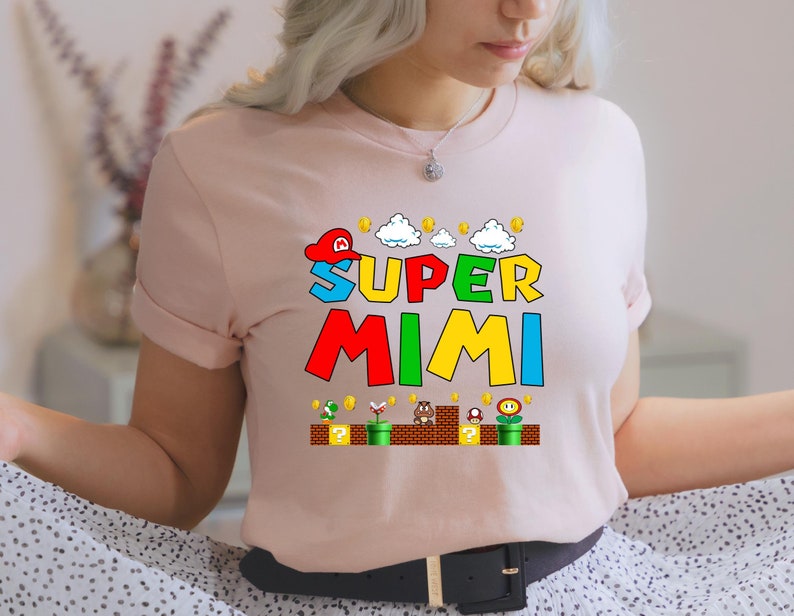 May include: A pink t-shirt with a graphic of a video game character wearing a red hat and the text "SUPER MIMI" in colorful letters. The graphic also includes a background of clouds, coins, and a brick platform with a question mark block.